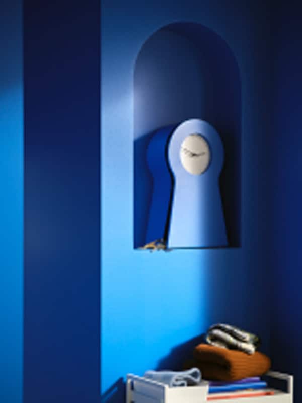 A blue IKEA PS 1995 clock with its storage cupboard open and a pair of keys beside it sitting on a blue shelf in a wall.
