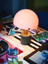 A light-pink-and-dark-grey-green NÖDMAST LED portable lamp lights up a tabletop covered in colourful textiles.