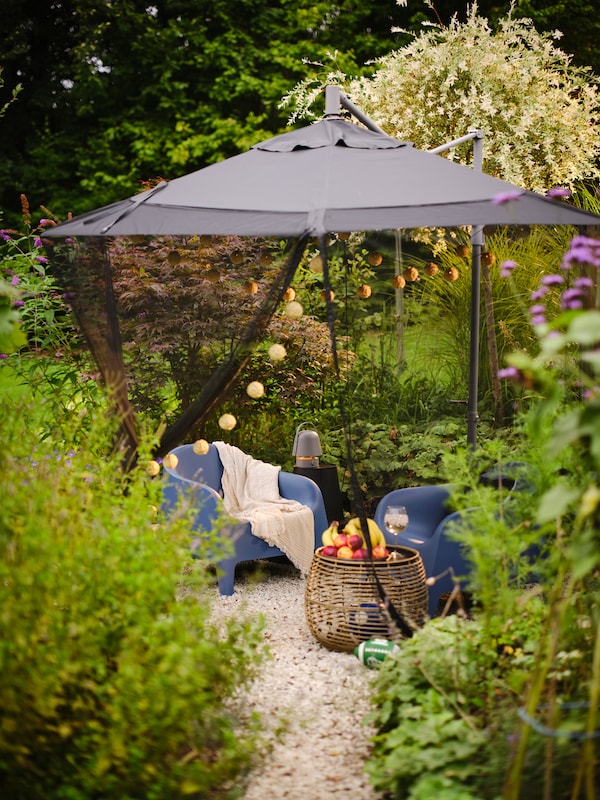 A lush garden with two dark blue SKARPÖ outdoor armchairs under a HISSÖ hanging parasol with a TORPÖN net hanging over it.