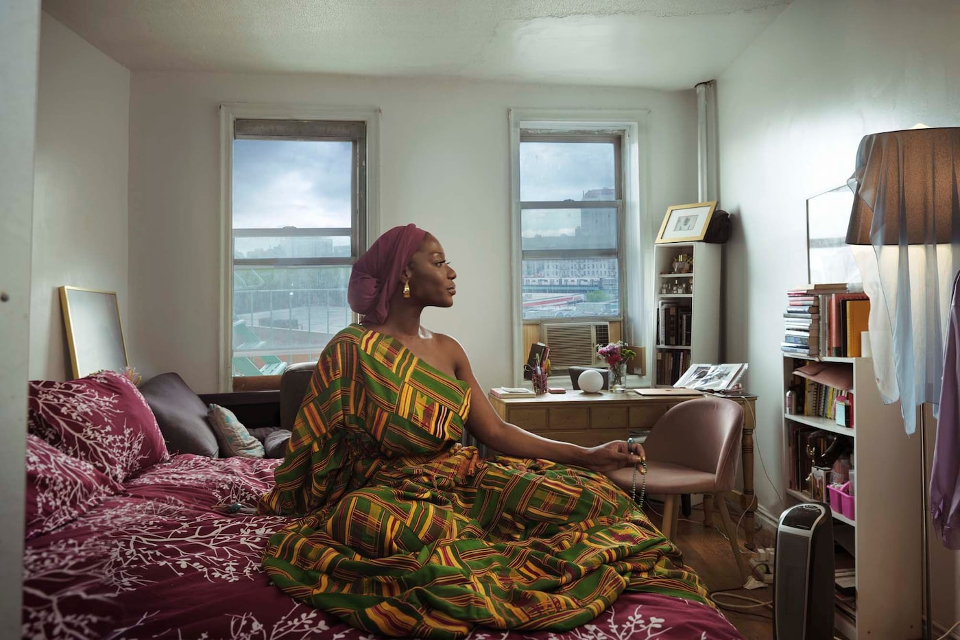 A woman wearing a brightly coloured patterned dress and headwrap sitting in an white-walled room. She is looking to the left, showing her face in profile. In the background there is a desk against a wall with two windows.