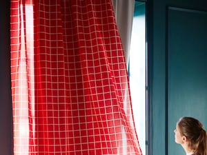 Una dona subjecta unes cortines vermelles mentre mira com el sol toca la finestra.