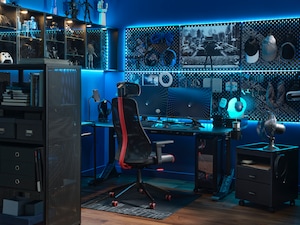 Gamerska soba, osvetljena z modro svetlobo, opremljena z dvema monitorjema, gaming stolom, pisalno mizo, omaricami, perforiranimi ploščami in vrsto gamerskih pripomočkov.