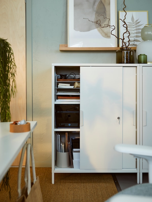 A light office interior with side-by-side white TROTTEN cabinets with sliding doors standing beneath framed art.