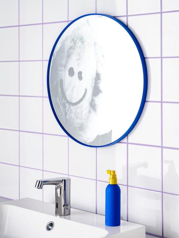 A blue LINDBYN mirror on a tiled wall and on top of a sink. A smiley face has been drawn on the mirror’s steamy surface.
