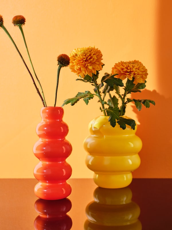 One orange and one yellow KÄLLARHALS vase with flowers in them are placed on a glossy surface that reflects the vases.