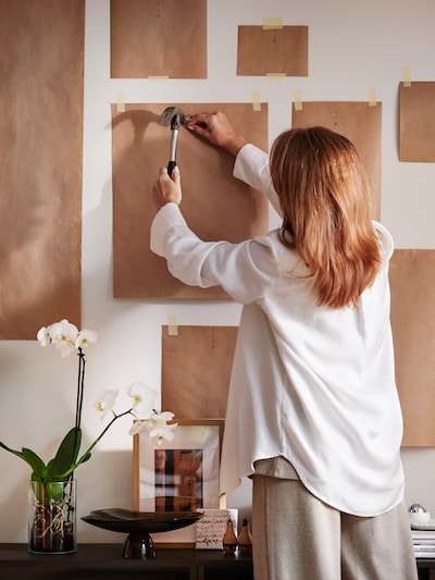A woman holds a hammer to a nail placed on one of many frame-sized paper templates taped to a wall in a bright living room.