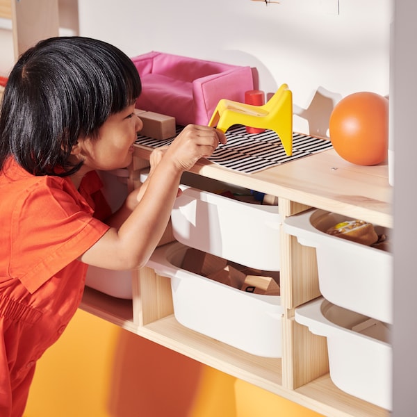A child picks a toy off the top of a light white stained pine TROFAST wall storage filled with white storage boxes.