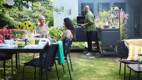 In a garden, a man uses the barbecue in a BÅTSKÄR outdoor kitchen while a family waits at a set TEGELÖN table for the food.