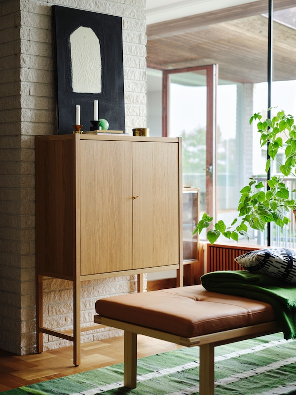 An oak veneer STOCKHOLM 2025 cabinet with two doors against a white brick wall, with a bench with a leather cushion in front.