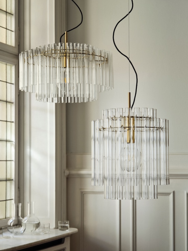 Two brass-plated STOCKHOLM 2025 pendant lamps with glass tubes side by side. Glasses and a carafe sit on a nearby windowsill.