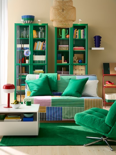 Living room with two green glass display cabinets side by side, small sofa-bed with multi-coloured cover and green rug.