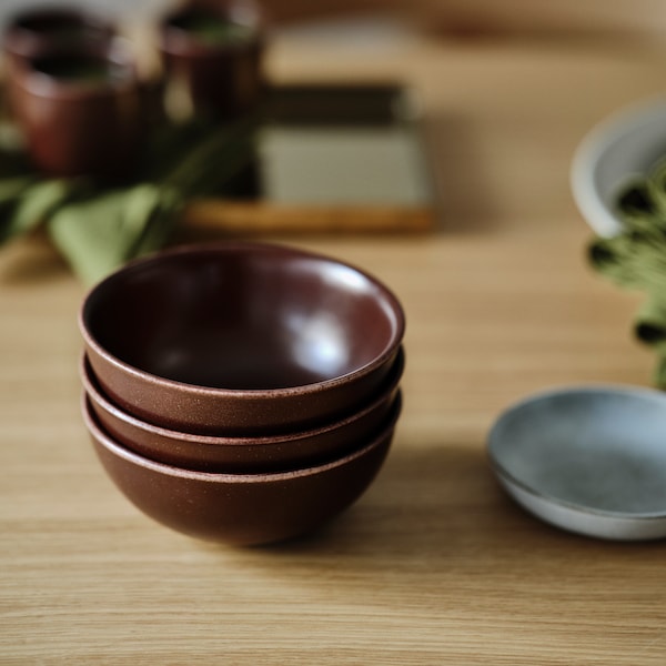 A tabletop with three stacked brown, Japanese-style STOCKHOLM 2025 bowls next to a rustic grey serving plate.