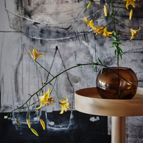 A brown STOCKHOLM 2025 glass vase with yellow flowers sitting on an oak veneer side table on castors.