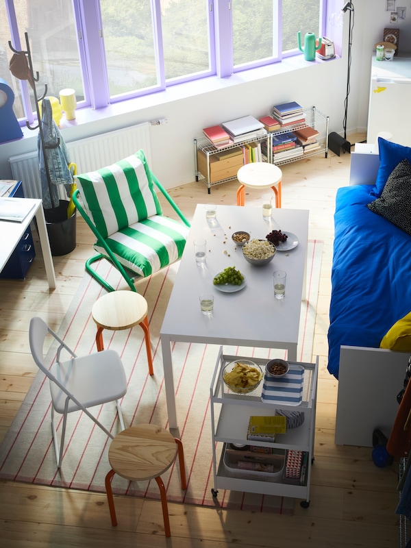 Picture taken from above, of a room with a day-bed and a table with different stools and chairs around. Big windows in back.