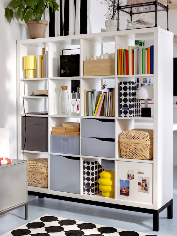 A white KALLAX shelving unit with many objects on its shelves including KALLAX inserts stands on a black KALLAX underframe.