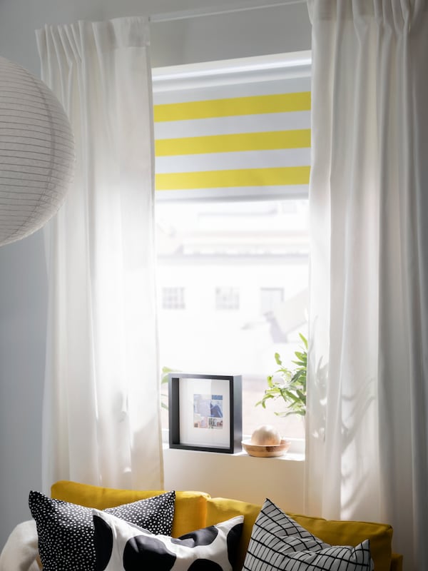 A striped FRIDANS block-out roller blind and GINSTMOTT curtains hang in a window above a yellow FRIDHULT sofa-bed.