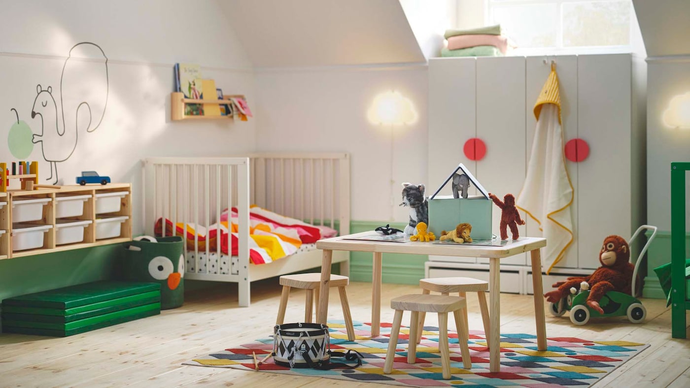 A todder bedroom with a table and chairs in the centre, a white cot in the left-hand corner and TROFAST storage on the wall.