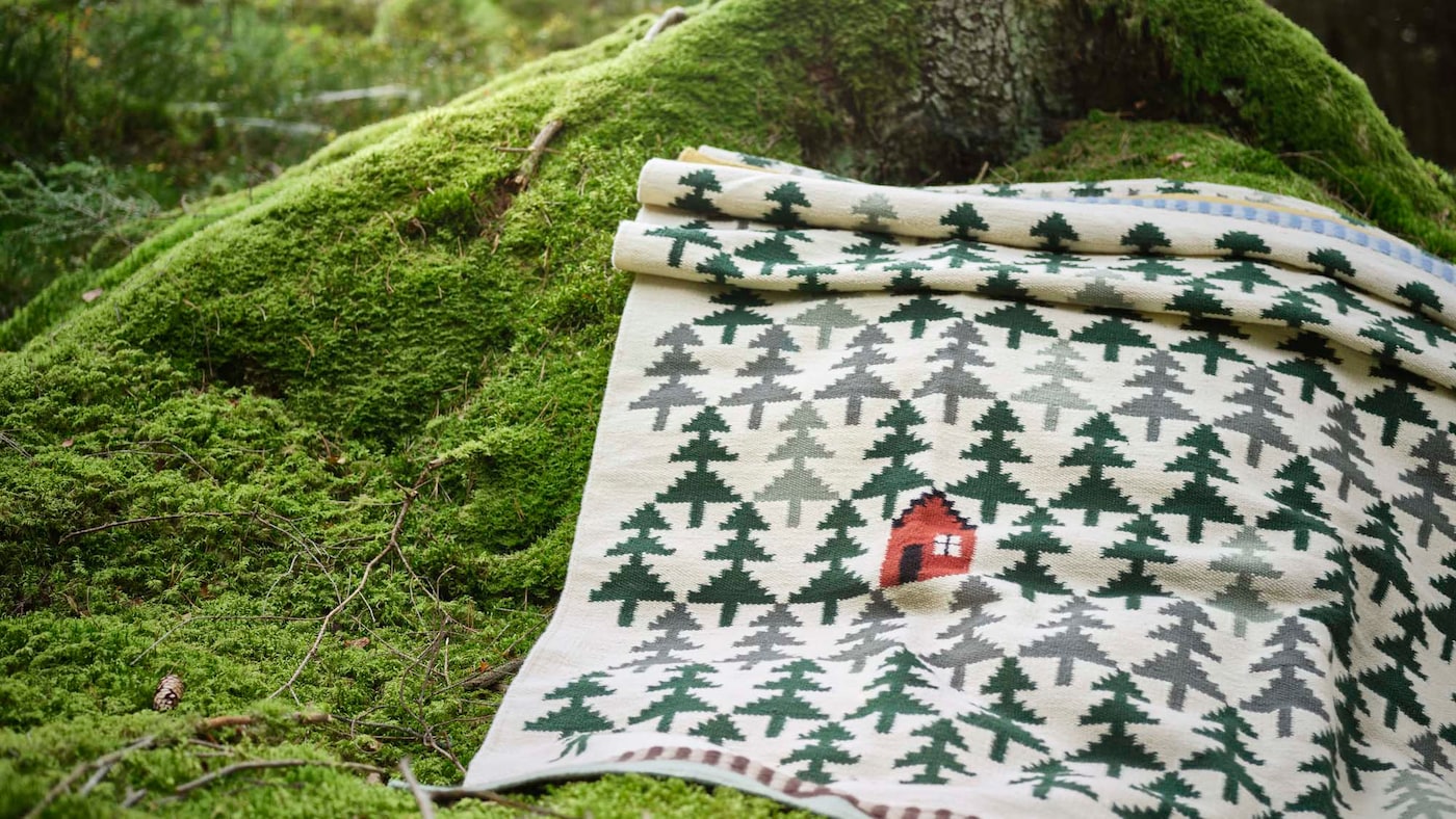 A DVÄRGGRÄS rug featuring a pine tree pattern with a little red house, is laid out on the mossy floor of a forest at the trunk of a tree.