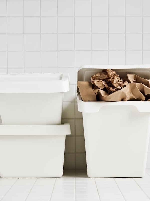 Tiga tong sampah SORTERA berwarna putih, salah satunya penuh dengan kertas berwarna coklat, terletak di ruang berjubin berwarna putih.